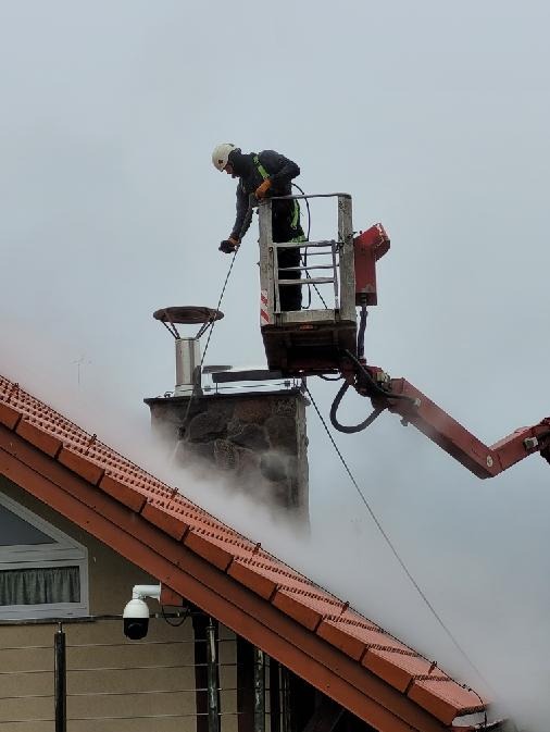 Pracownik w kasku na podnośniku koszowym czyści komin na dachu budynku pokrytego czerwoną dachówką, w tle widoczny dom z kamerą monitoringu.