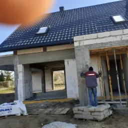 Budowa domu w stanie surowym otwartym z dachem krytym czarną dachówką, widoczne białe bloczki Solbet i pracownik w kurtce z napisem RK Niedziałek przy ścianie.