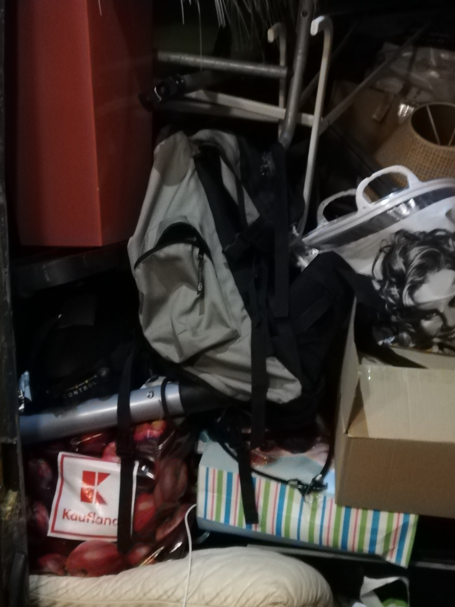 Stłoczone przedmioty w ciemnym pomieszczeniu, w tym plecak, drabina aluminiowa, torba z logiem Kaufland i kartonowe pudełko.