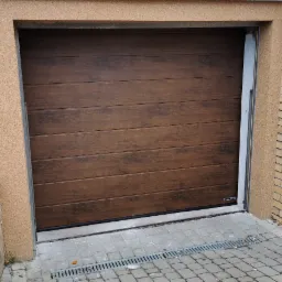 Zamknięta, segmentowa brama garażowa w odcieniach brązu z poziomymi panelami, widoczny fragment elewacji budynku z fakturą baranka oraz kostka brukowa przed wjazdem.