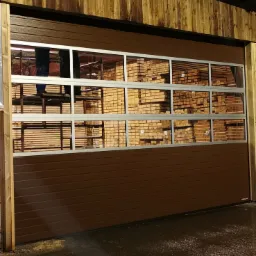 Brązowa segmentowa brama z oknami, za którą widoczne składowane drewno, częściowo zasłonięte przez elementy rusztowania i postać w roboczych ubraniach.