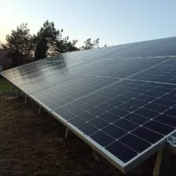 Rząd paneli słonecznych na metalowej konstrukcji, zainstalowany na ziemi obok budynku z widocznymi drzewami w tle, sfotografowany w pochmurny dzień.