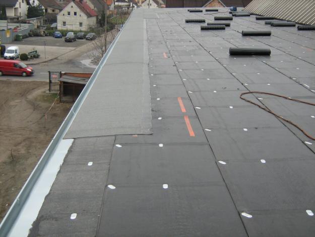 Widok z góry na płaski dach pokryty czarną papą termozgrzewalną, z widocznymi połączeniami i elementami mocującymi, częściowo wykończony szarym pasem papy przy krawędzi z orynnowaniem, na tle...