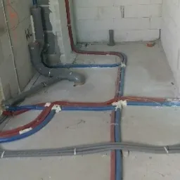 Instalacja wodna i kanalizacyjna w surowym stanie, rury w kolorach niebieskim, czerwonym i szarym, zabezpieczone pianką montażową, widoczne ściany z bloczków.