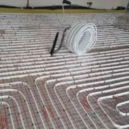 Białe rury ogrzewania podłogowego ułożone w pętle na podkładzie z siatką w nowym budynku, widoczne narzędzie do rozwijania rur.