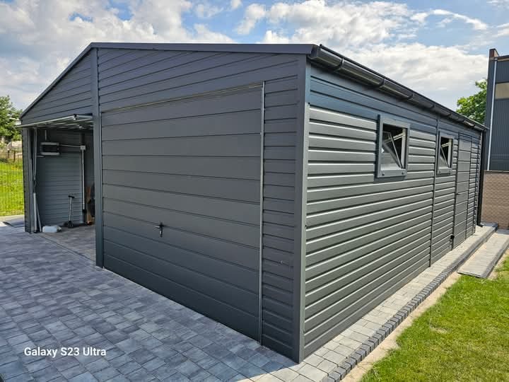 Nowoczesny, grafitowy garaż blaszany z dachem dwuspadowym, widoczne okna i brama segmentowa, położony na kostce brukowej w słoneczny dzień.