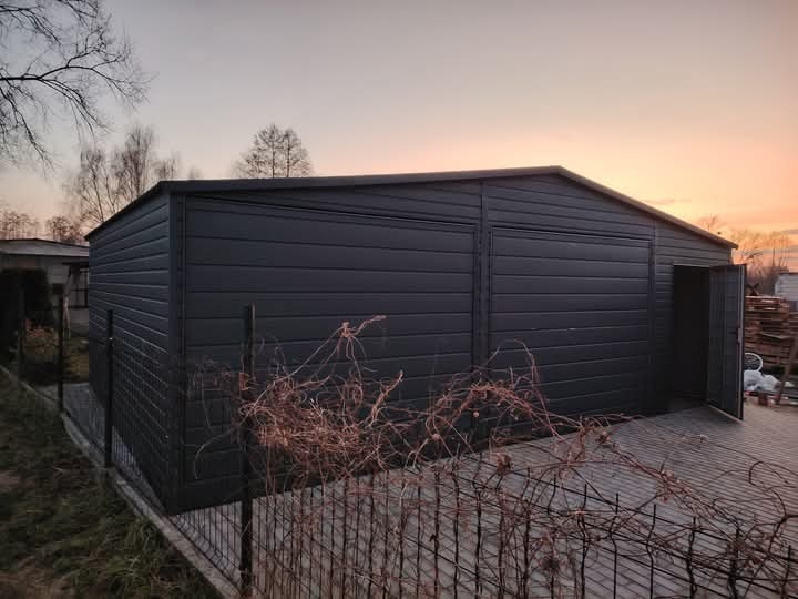 Nowoczesny, antracytowy garaż dwustanowiskowy z blachy trapezowej, z otwartymi drzwiami po prawej stronie, na posesji z betonową kostką, widok o zmierzchu.
