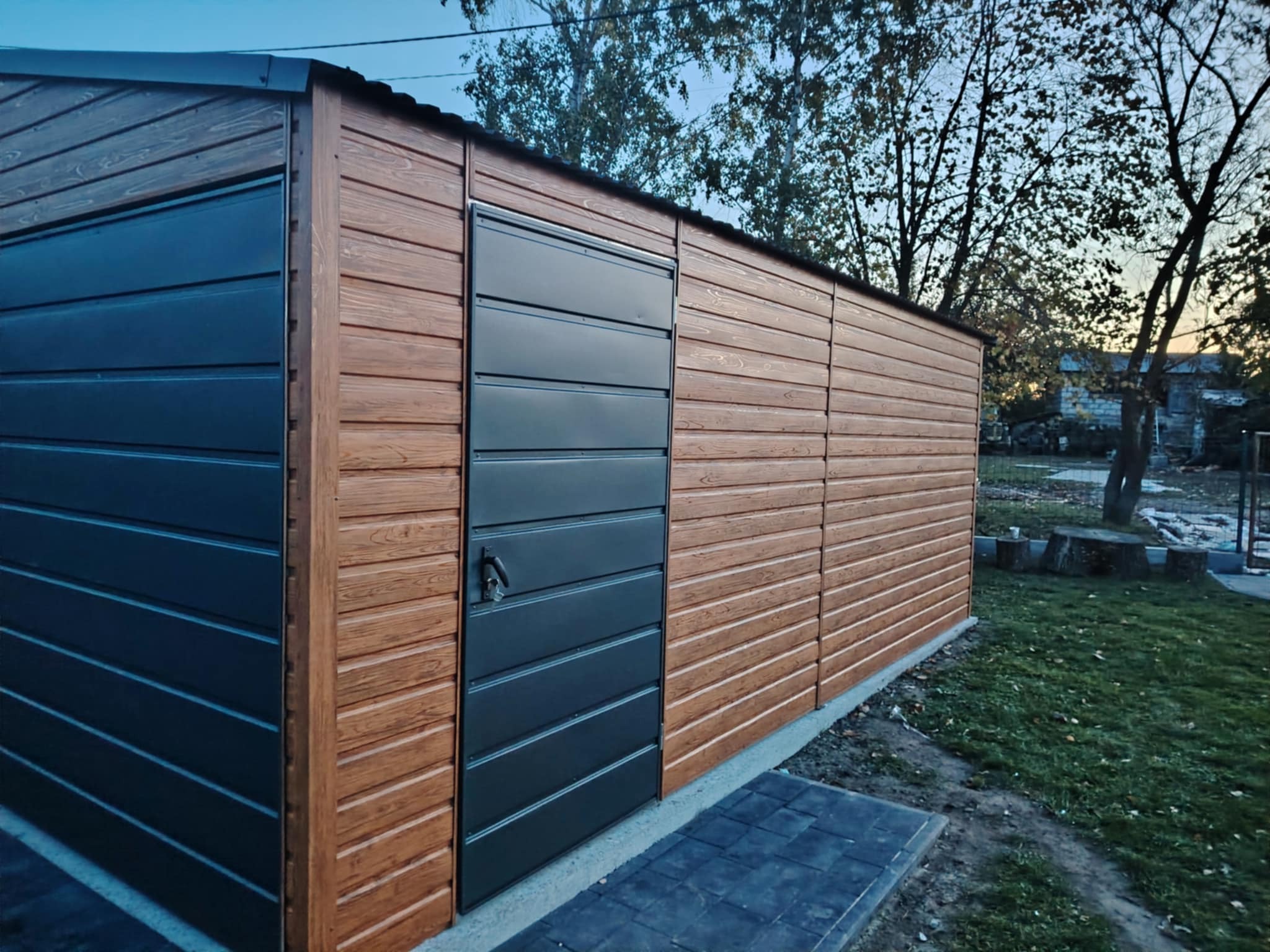 Nowoczesny garaż blaszany z drewnopodobną elewacją i antracytowymi drzwiami segmentowymi, widok z boku na tle zielonego trawnika i drzew.