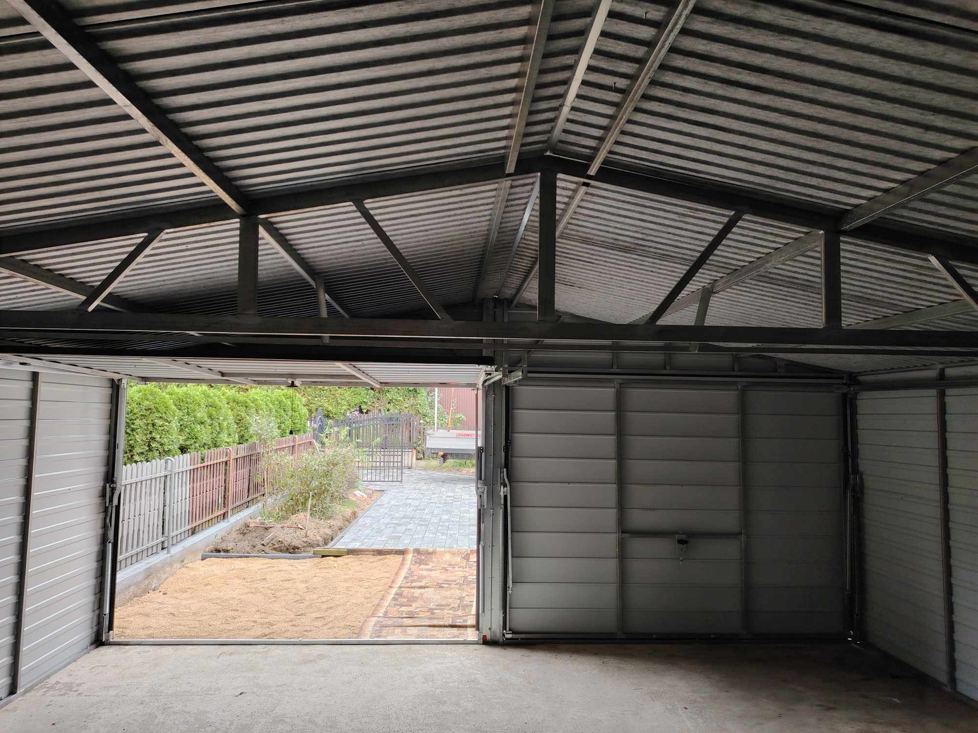 Wnętrze blaszanego garażu z otwartymi drzwiami, widok na posesję z kostką brukową i ogrodzeniem, konstrukcja dachu z blachy falistej.