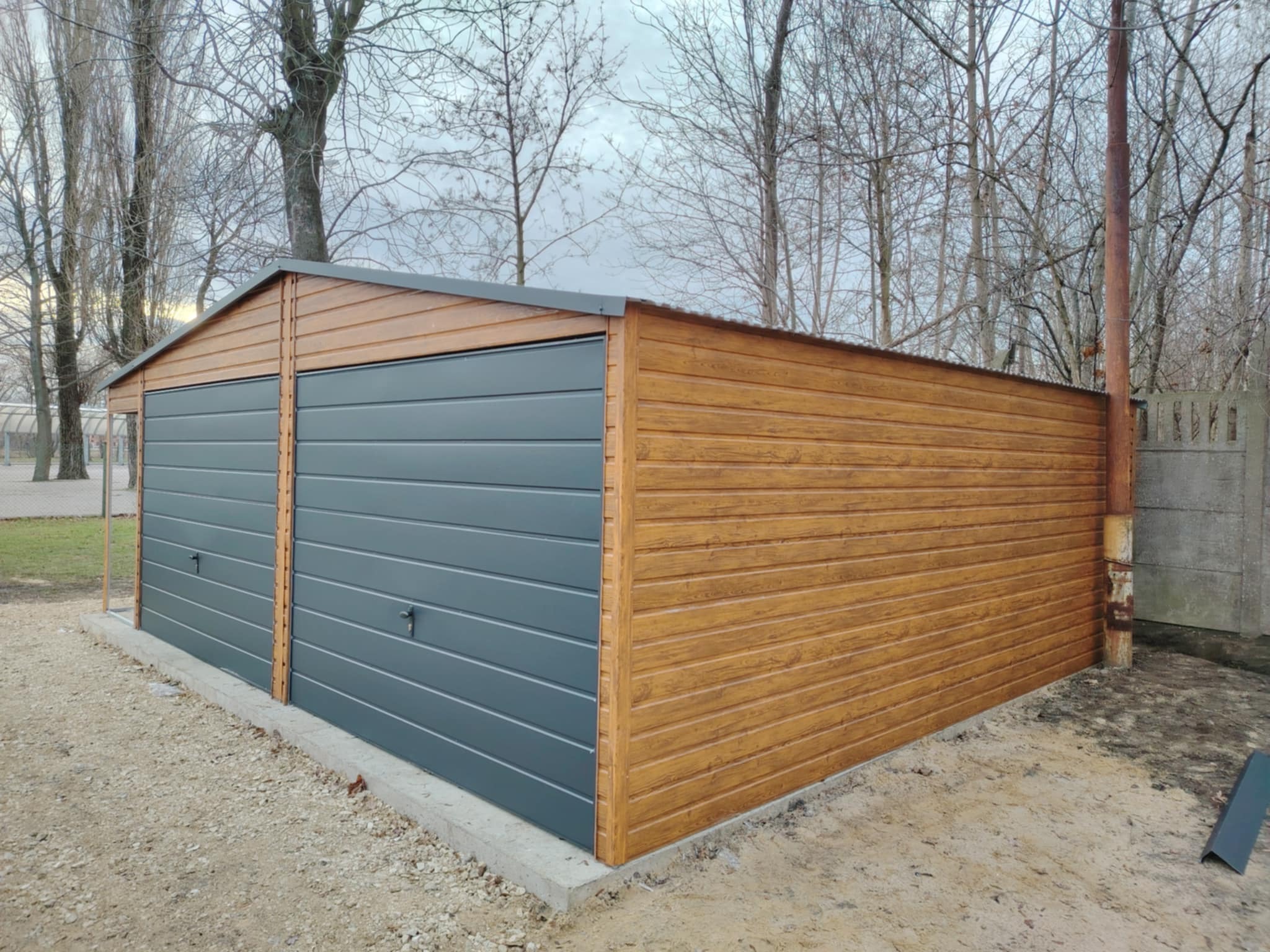 Dwustanowiskowy garaż z blachy trapezowej, z dwiema szarymi bramami segmentowymi i drewnopodobną elewacją, na betonowej wylewce, widok z zewnątrz w pochmurny dzień.