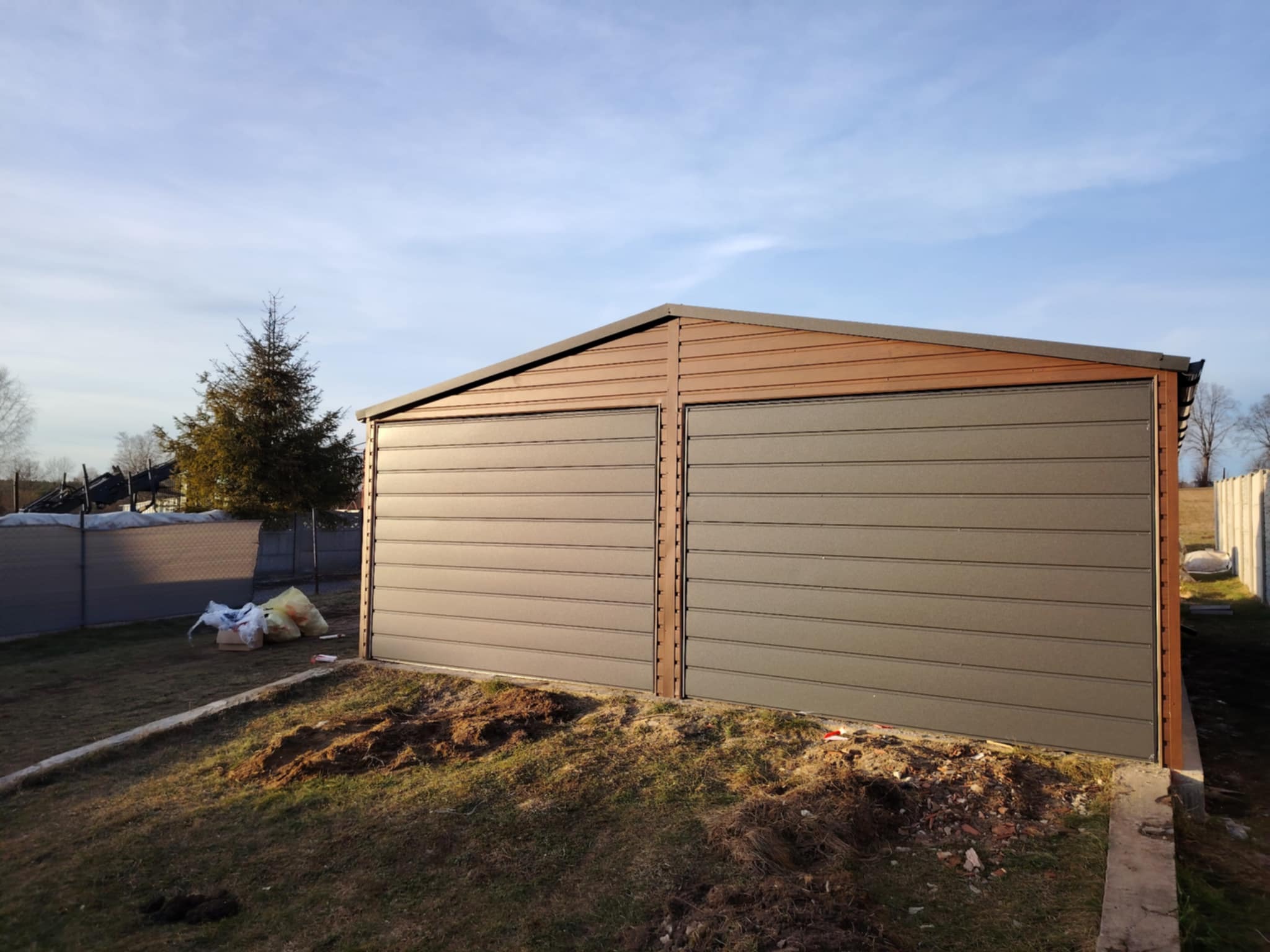 Nowoczesny, dwustanowiskowy garaż blaszany z brązowym wykończeniem dachu i szarymi panelami, ustawiony na nierównym terenie trawiastym z widocznymi workami z materiałami budowlanymi obok.