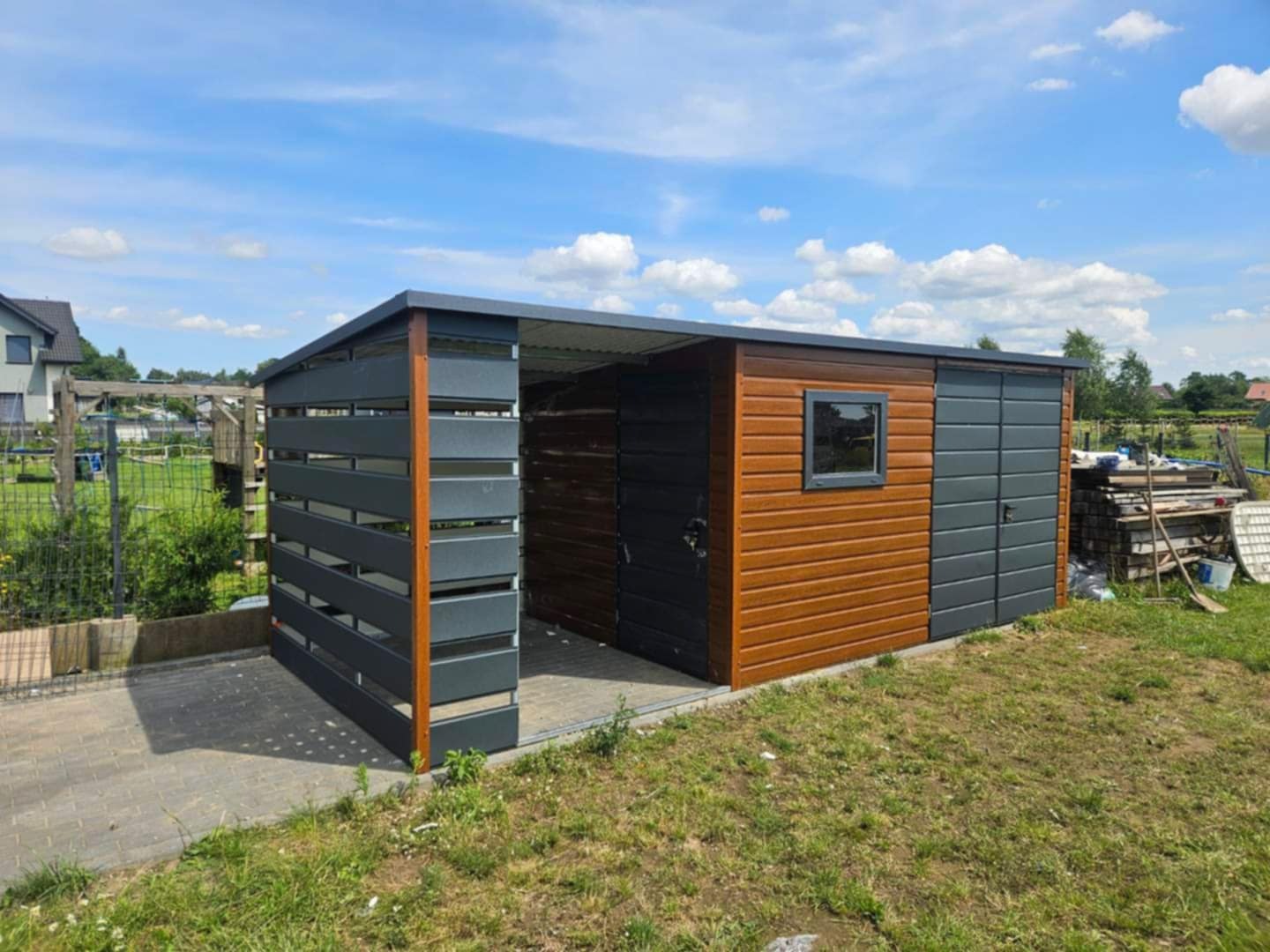 Nowoczesny garaż blaszany z elementami drewnopodobnymi i ażurową ścianą boczną, widoczny na tle zielonej trawy i błękitnego nieba.