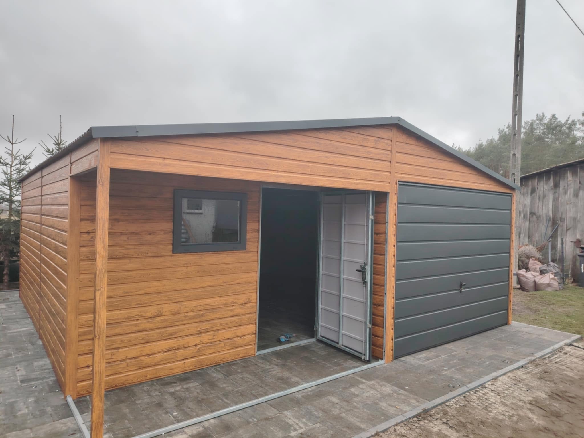 Nowoczesny garaż blaszany z drewnopodobną elewacją, szarą bramą segmentową i otwartymi dwuskrzydłowymi drzwiami wejściowymi, usytuowany na posesji z betonową kostką brukową.