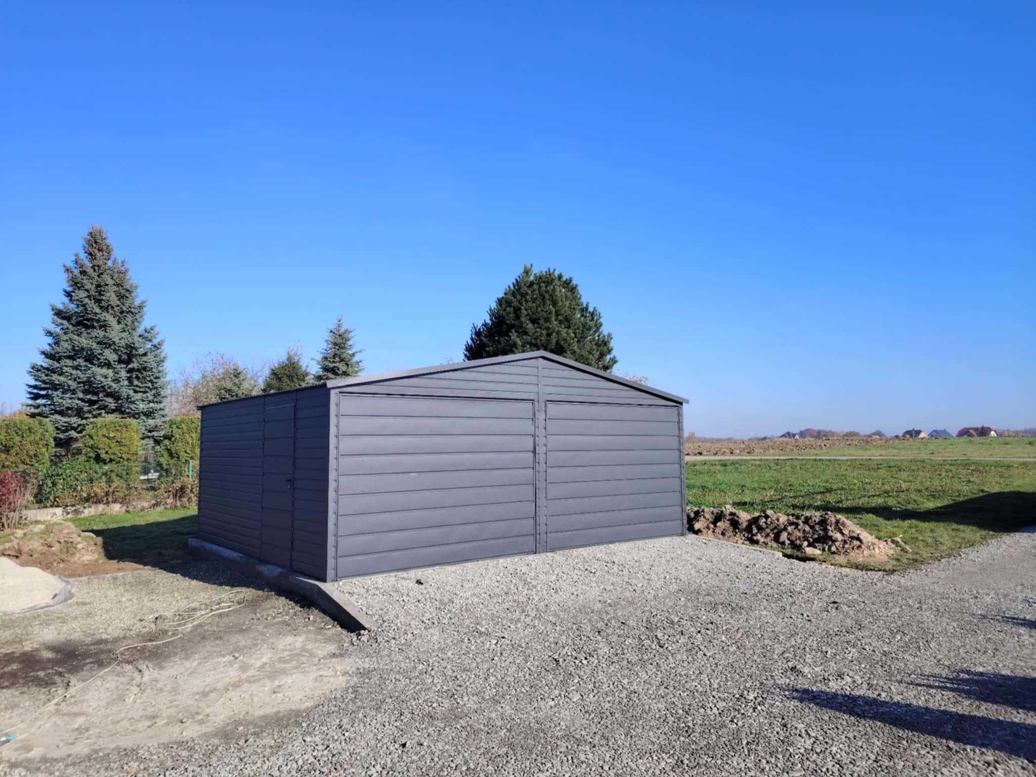 Szary garaż blaszany dwustanowiskowy z dwuskrzydłowymi drzwiami, ustawiony na wysypanym żwirem podłożu, widok z boku z perspektywy gruntu, w tle zieleń i błękitne niebo.