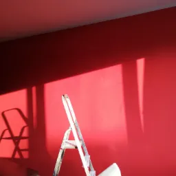 Świeżo pomalowana na czerwono ściana w pokoju oświetlona popołudniowym słońcem, z metalową drabiną opartą o ścianę i fragmentem krzesła w kadrze.