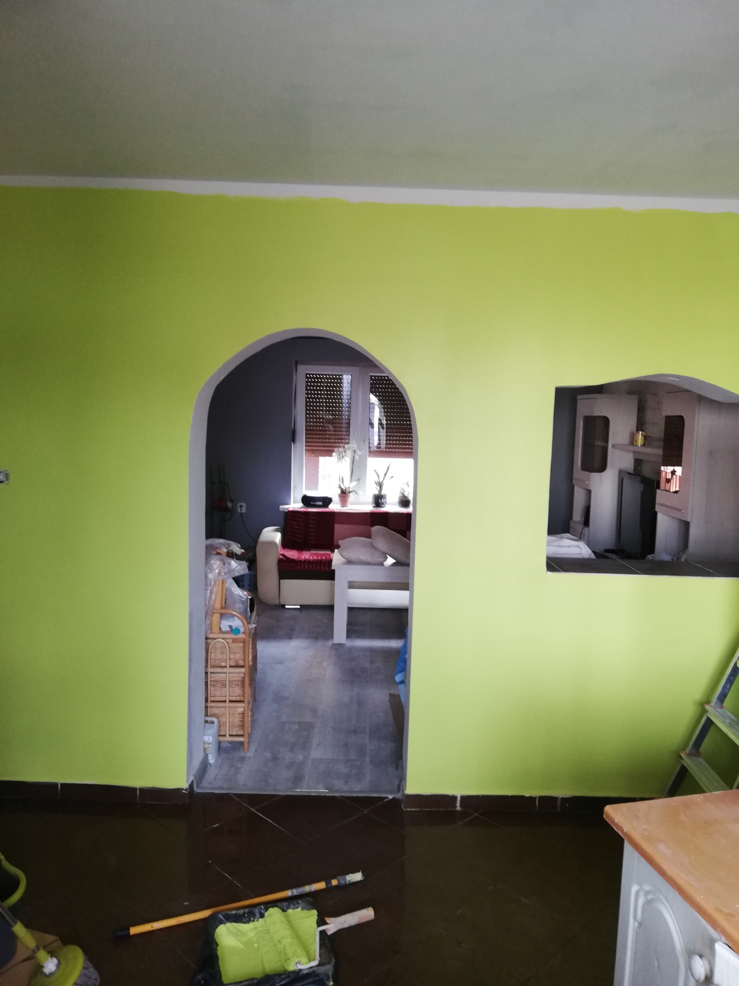 Świeżo pomalowane na limonkowo ściany z widocznymi łukowatymi przejściami do innych pomieszczeń, wałek i kuweta z farbą na podłodze, widoczny fragment drabiny.