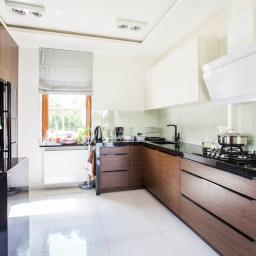 M-Bud - Nowoczesna kuchnia z drewnianymi szafkami dolnymi i białymi górnymi, czarnym blatem, białymi płytkami podłogowymi i oknem z roletą rzymską, częściowo widoczny sprzęt AGD.