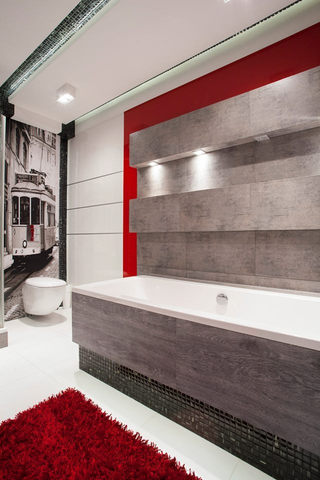 Nowoczesna łazienka z wanną obudowaną panelami imitującymi drewno, szara ściana z podświetlanymi półkami i czerwonym akcentem, fototapeta z tramwajem oraz czerwony dywanik na białej podłodze.