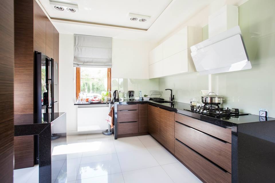 Nowoczesna kuchnia z drewnianymi szafkami dolnymi i białymi górnymi, czarnym blatem, białymi płytkami podłogowymi i oknem z roletą rzymską, częściowo widoczny sprzęt AGD.