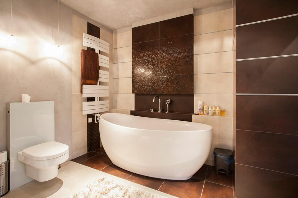Nowoczesna łazienka z wolnostojącą wanną, brązowymi płytkami imitującymi skórę na ścianie i białym grzejnikiem drabinkowym z ręcznikiem.