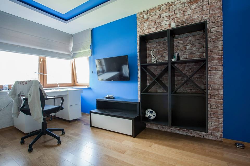 Pokój młodzieżowy z niebieską ścianą, ceglaną wstawką i czarnymi regałami, drewniana podłoga, biurko przy oknie z roletami rzymskimi.