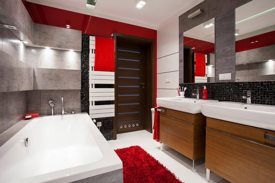 Nowoczesna łazienka z białą wanną, dwiema umywalkami na drewnianych szafkach, czerwonym akcentem na suficie i dywaniku oraz grzejnikiem drabinkowym.