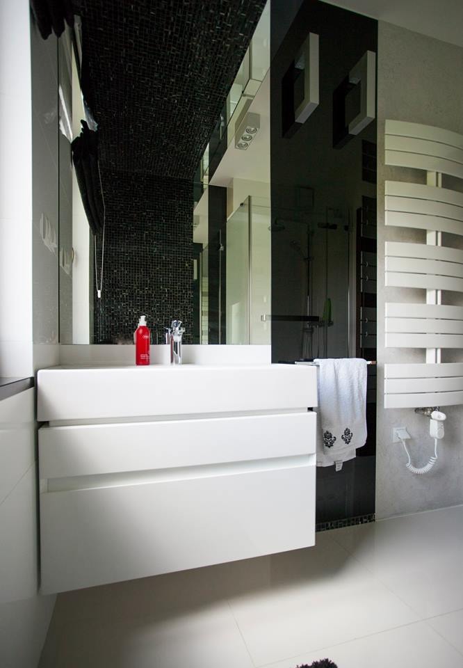 Nowoczesna łazienka w czerni i bieli: biała umywalka z szafką, czarny panel szklany, grzejnik drabinkowy, czarna mozaika na suficie i kabina prysznicowa w tle.