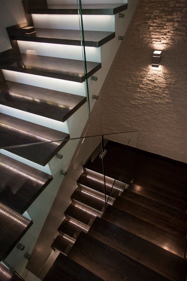 Nowoczesne schody z ciemnego drewna z podświetleniem LED na stopniach i szklaną balustradą, oświetlone kinkietem na dekoracyjnej ścianie z kamienia.