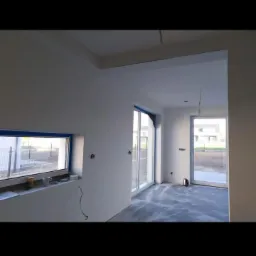 Wnętrze domu w trakcie prac wykończeniowych: świeżo pomalowane ściany na biało, widoczne okno z niebieską ramą i przesuwne drzwi tarasowe, surowa podłoga przygotowana do dalszych prac.