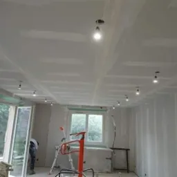 Pokój w trakcie szpachlowania: ściany i sufit pokryte gładzią gipsową, widoczne punkty oświetleniowe w suficie podwieszanym, drabina i narzędzia malarskie w tle.