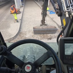 WASILJEW - Widok z kabiny koparki na plac budowy: robotnik z łopatą, przygotowany teren pod kostkę i ramię koparki. Praca wre, a operator ma wszystko pod kontrolą.