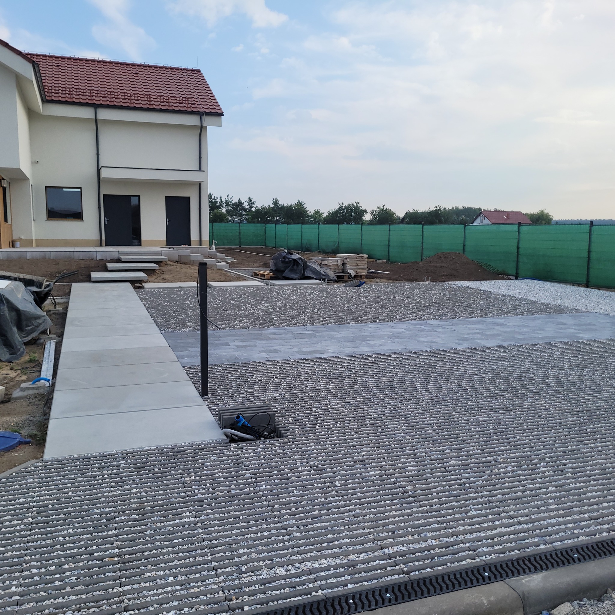 Niedokończona nawierzchnia z ażurowych płyt betonowych wypełnionych drobnym kamieniem, z widoczną studzienką kanalizacyjną, fragmentem chodnika z płyt i nowym domem w tle, zabezpieczonym zielonym...