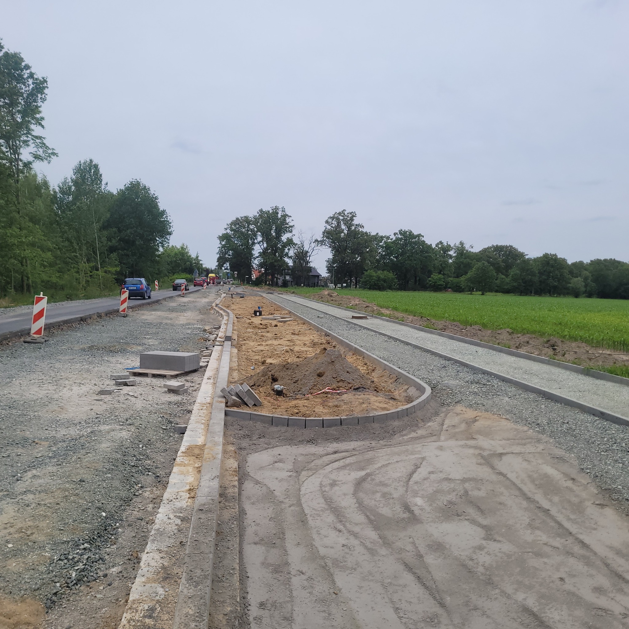 Budowa drogi z obrzeżami i podsypką, widoczne przygotowane miejsce pod kostkę, w tle pole i drzewa, na drodze w oddali samochody.