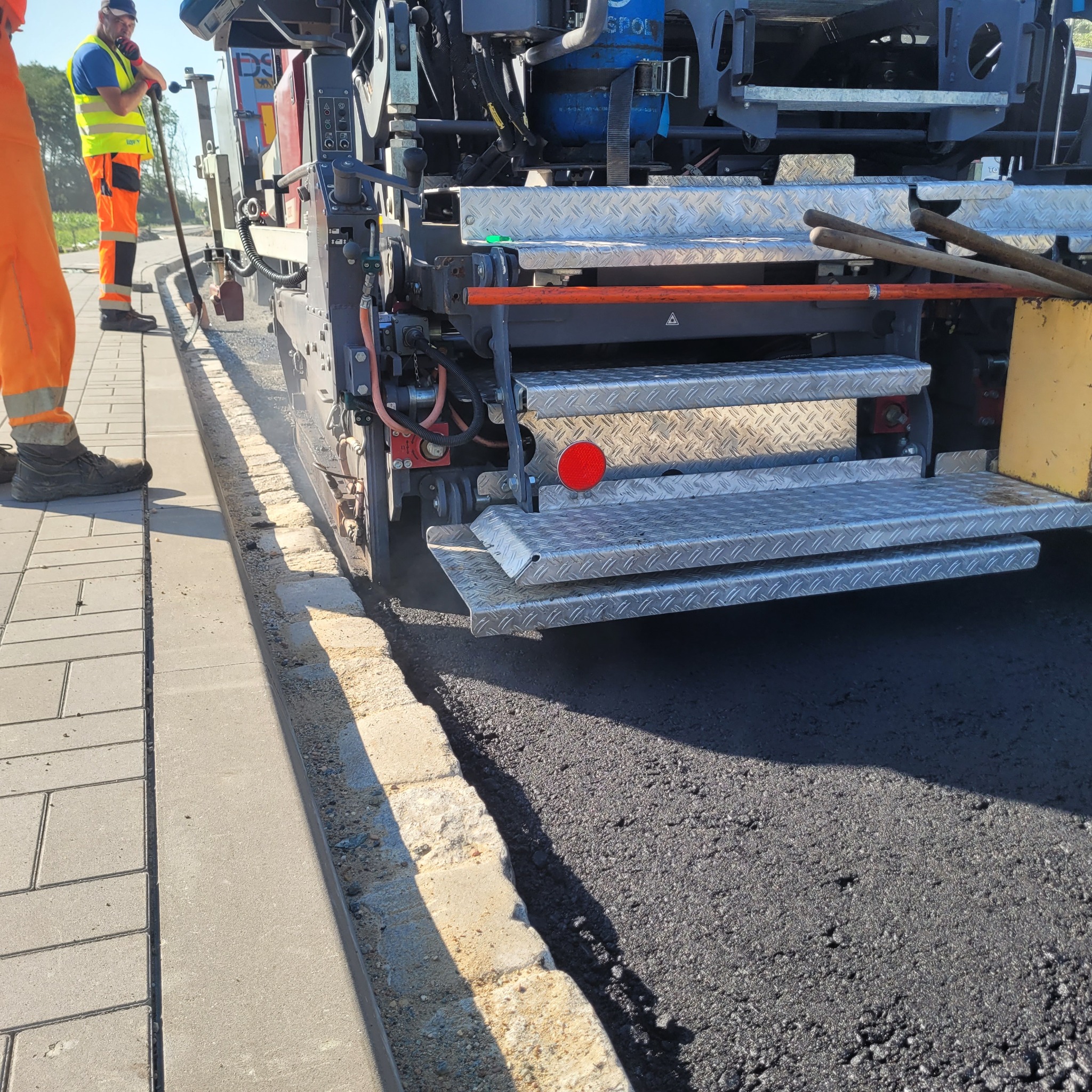 Układanie asfaltu przy krawężniku z kostki brukowej za pomocą rozściełacza; widoczni pracownicy w odblaskowych ubraniach.