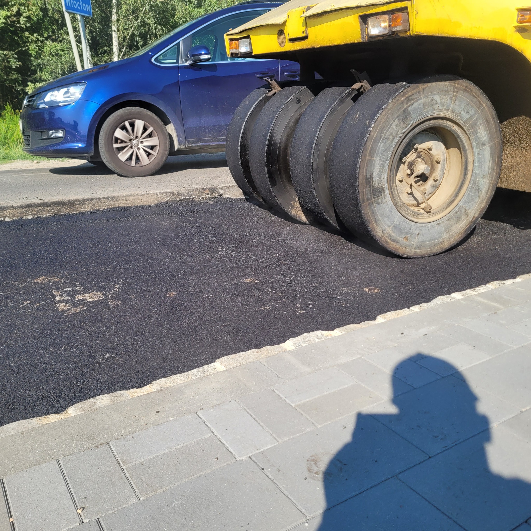 Walec drogowy ugniata świeżo wylany asfalt w Radwanicach, na tle zaparkowanego niebieskiego samochodu osobowego.