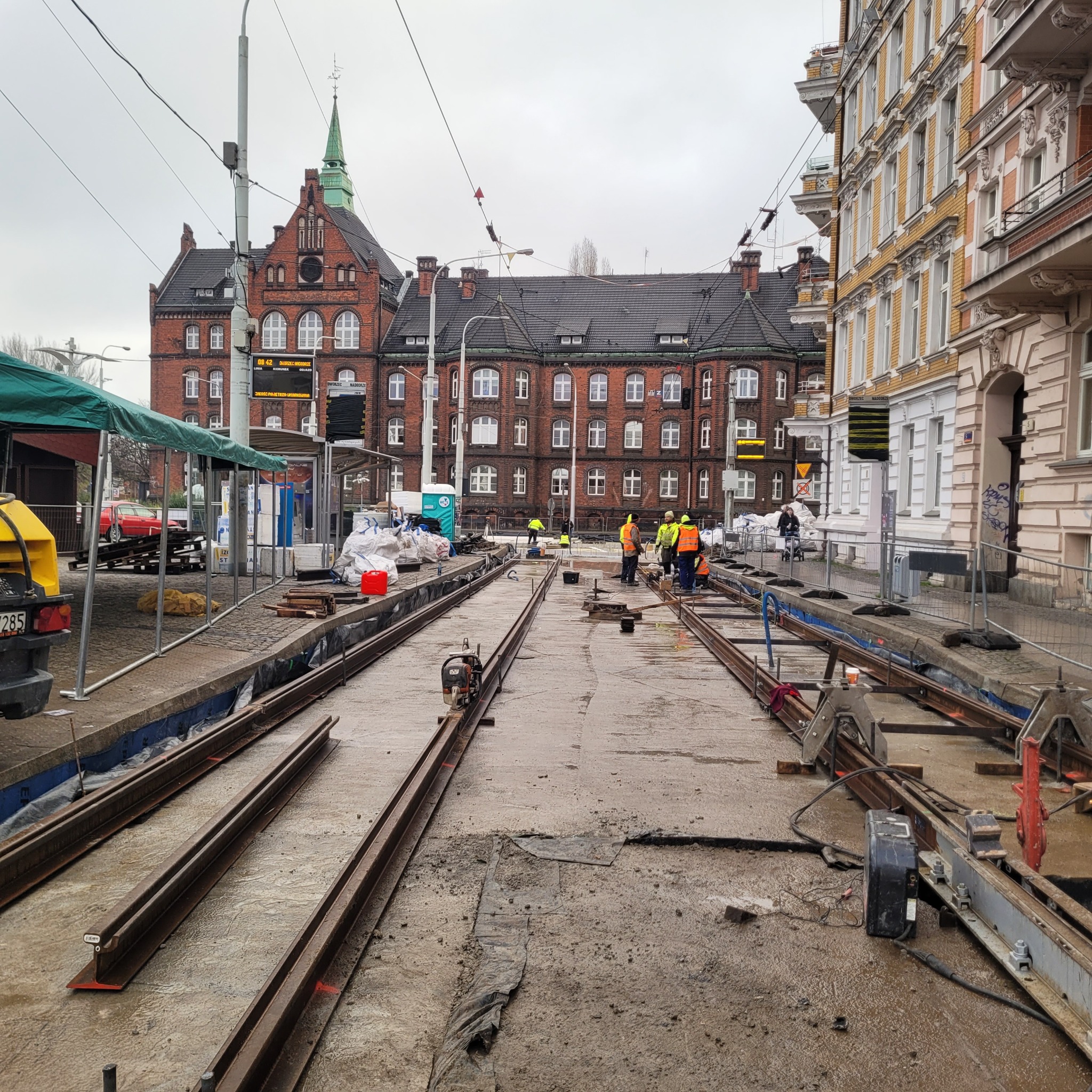 Montaż torów tramwajowych na tle miejskiej zabudowy, robotnicy w odblaskowych kamizelkach przy pracy, widoczne narzędzia i materiały budowlane, pochmurne niebo.