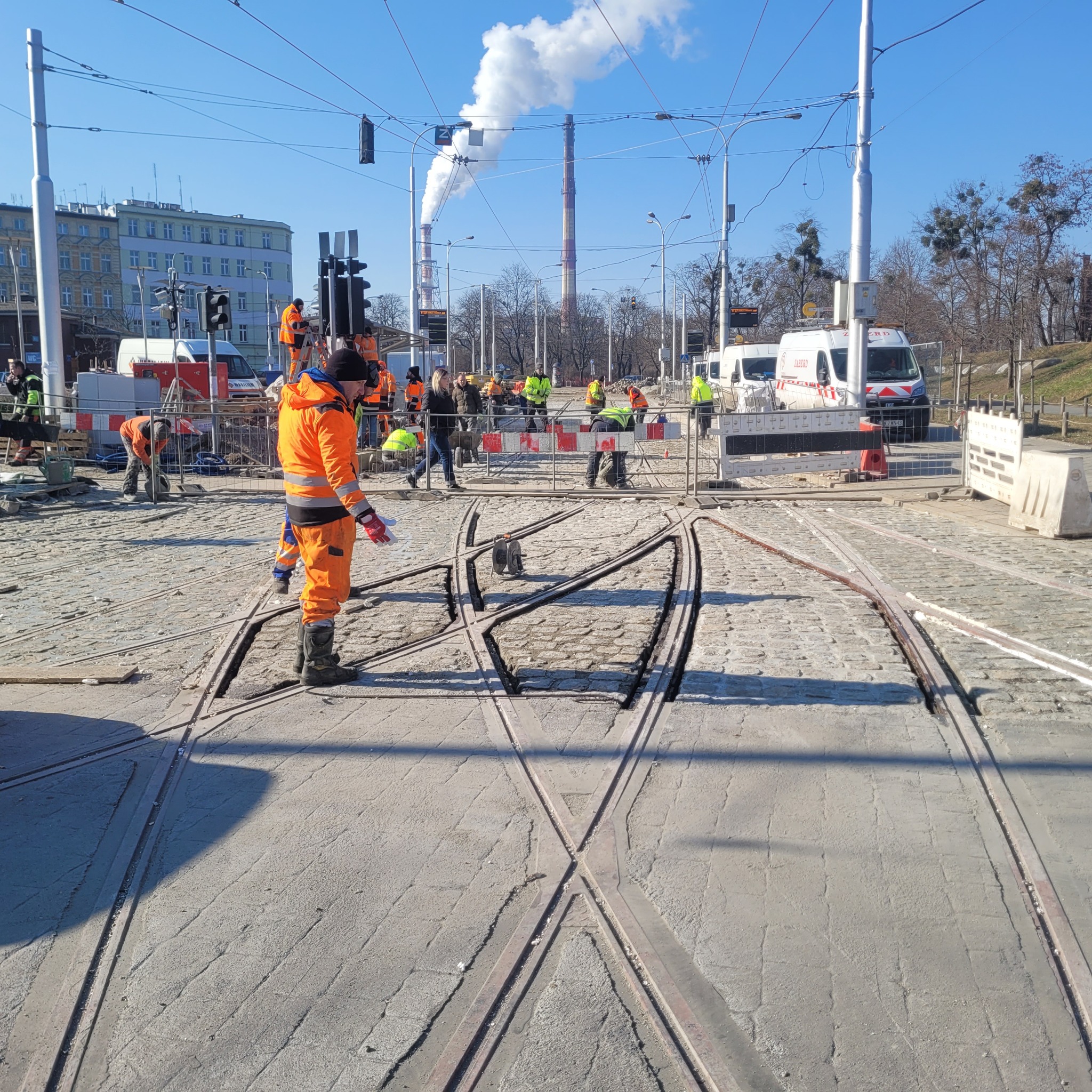 Prace brukarskie przy torach tramwajowych w mieście, robotnicy w pomarańczowych kombinezonach, widoczne fragmenty kostki brukowej i elementy torowiska, w tle budynki i kominy.
