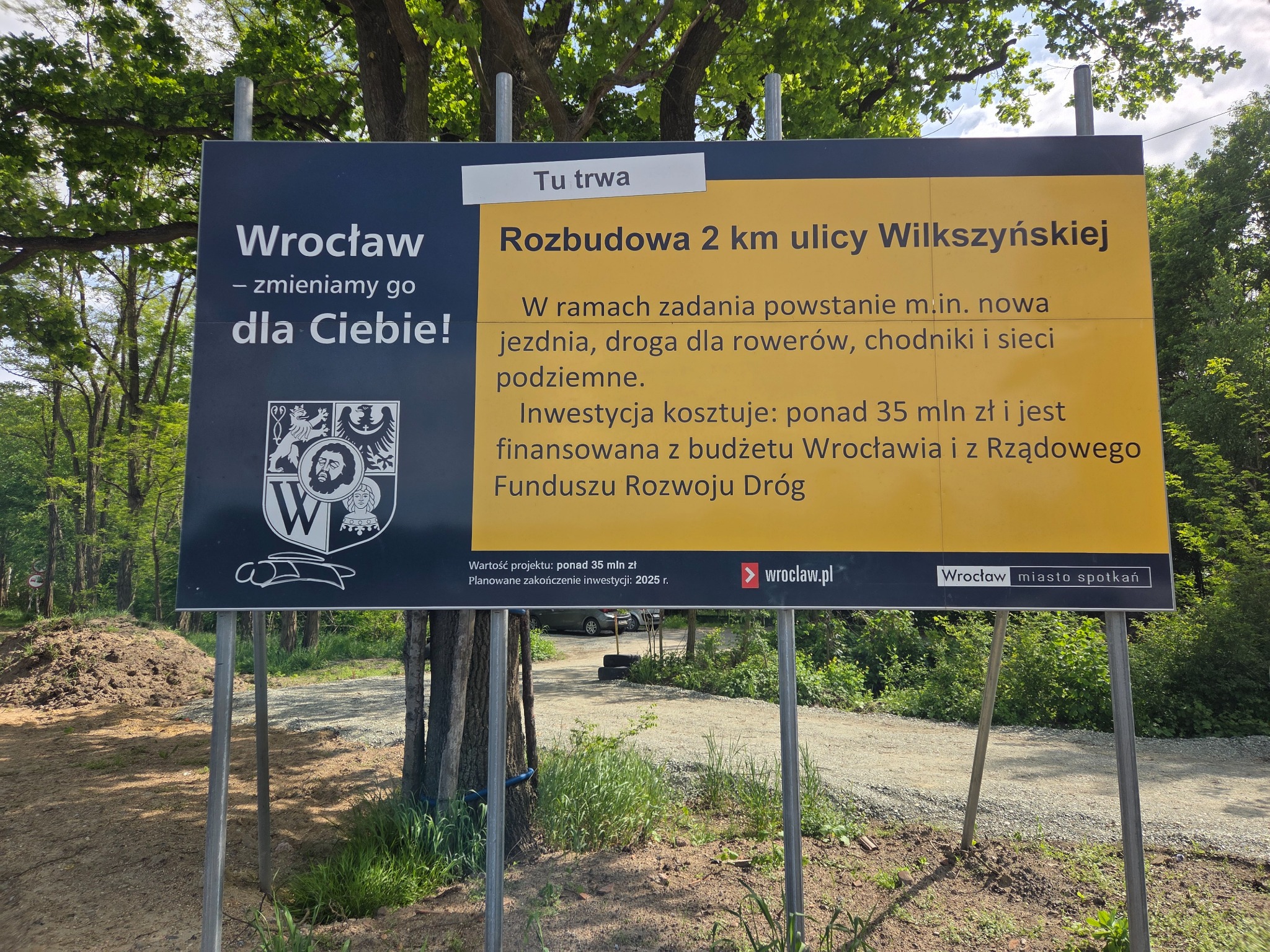 Tablica informacyjna o rozbudowie ulicy Wilkszyńskiej we Wrocławiu, z herbem miasta i informacjami o inwestycji.