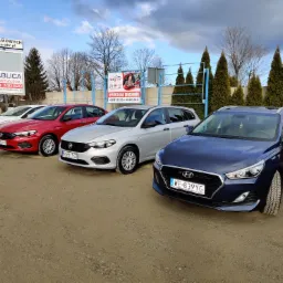Rząd zaparkowanych samochodów różnych marek i kolorów na żwirowym parkingu, z reklamą firmy z branży motoryzacyjnej w tle.