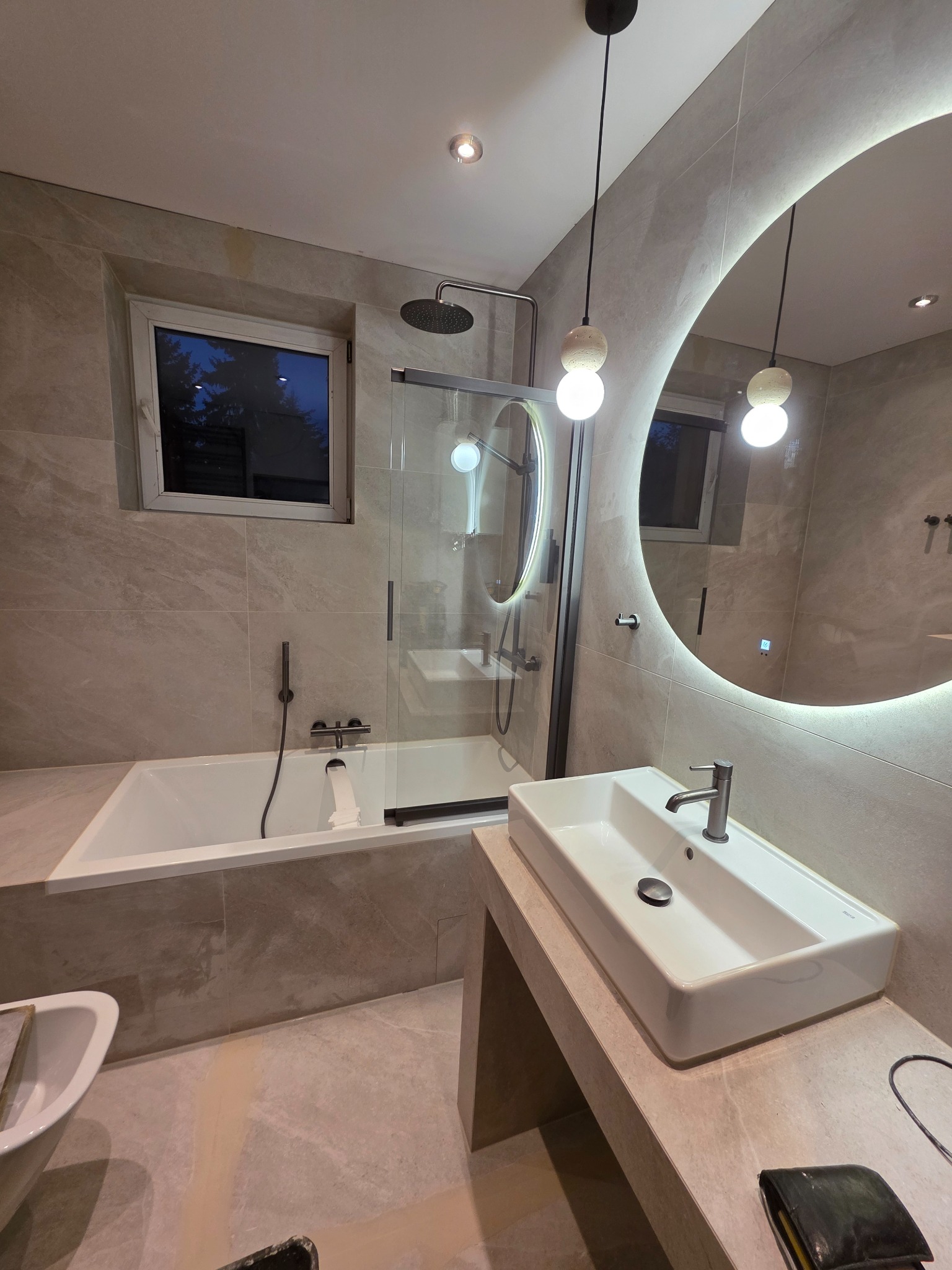 Wykończona łazienka w beżowych płytkach z wanną, kabiną prysznicową, umywalką i okrągłym lustrem z podświetleniem LED. Widoczne narzędzia glazurnicze.