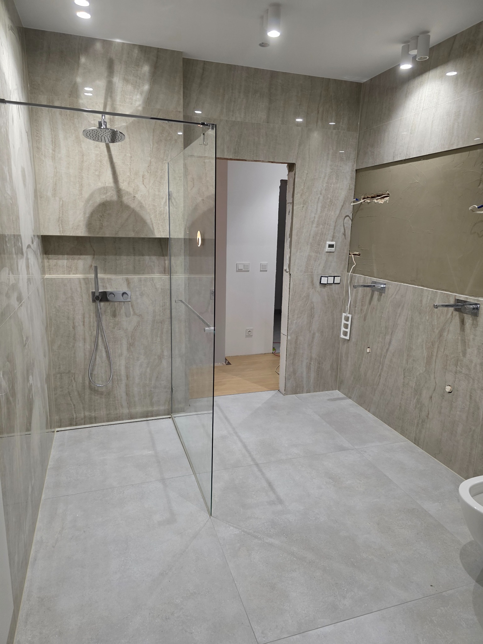 Wykończona łazienka z kabiną prysznicową typu walk-in, płytki imitujące kamień na ścianach i podłodze, widoczne elementy instalacji elektrycznej i hydraulicznej.