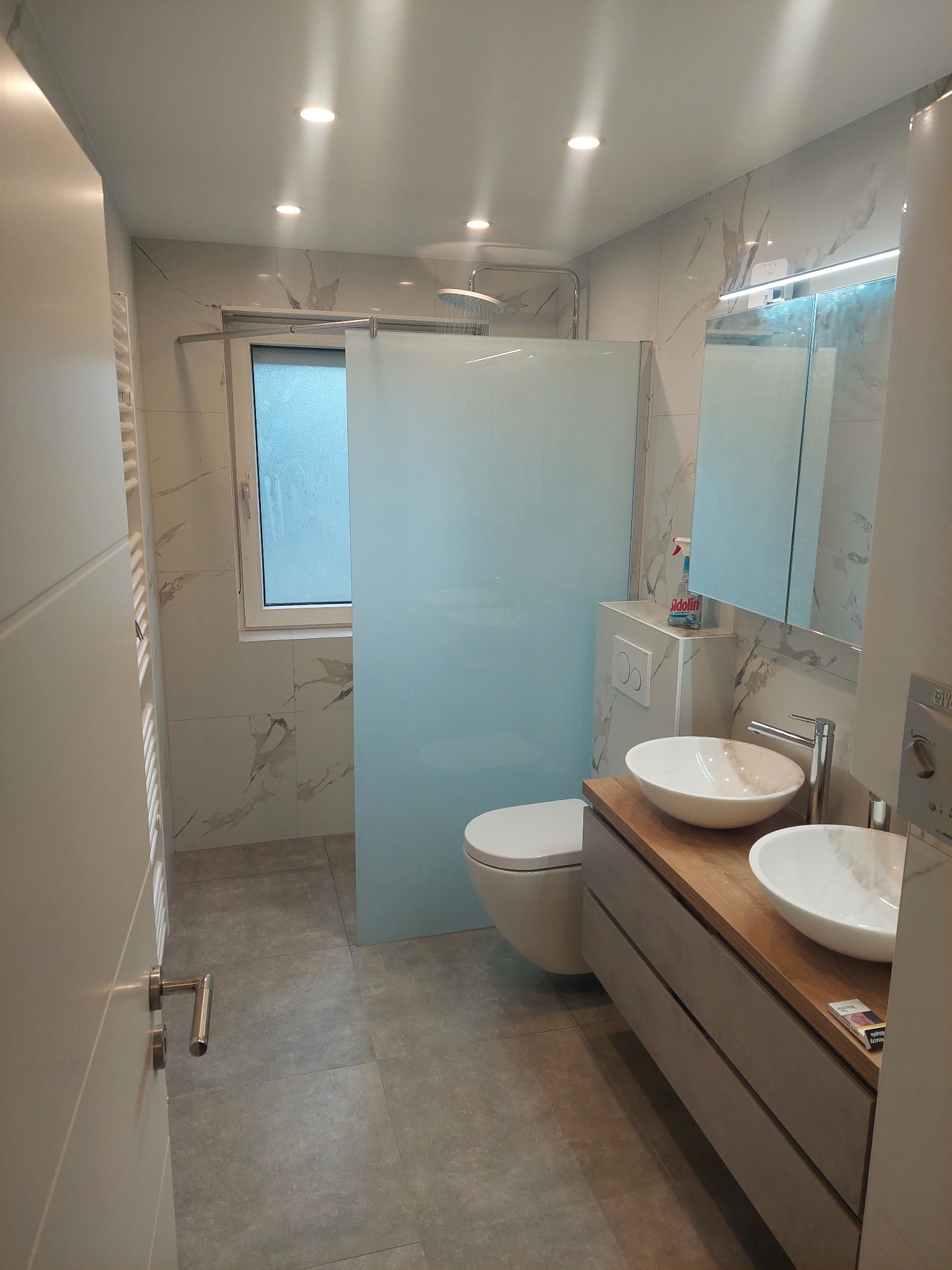 Nowoczesna łazienka po remoncie: marmurowe płytki, szklana kabina prysznicowa, dwie umywalki nablatowe na drewnianym blacie, szara podłoga i okno.