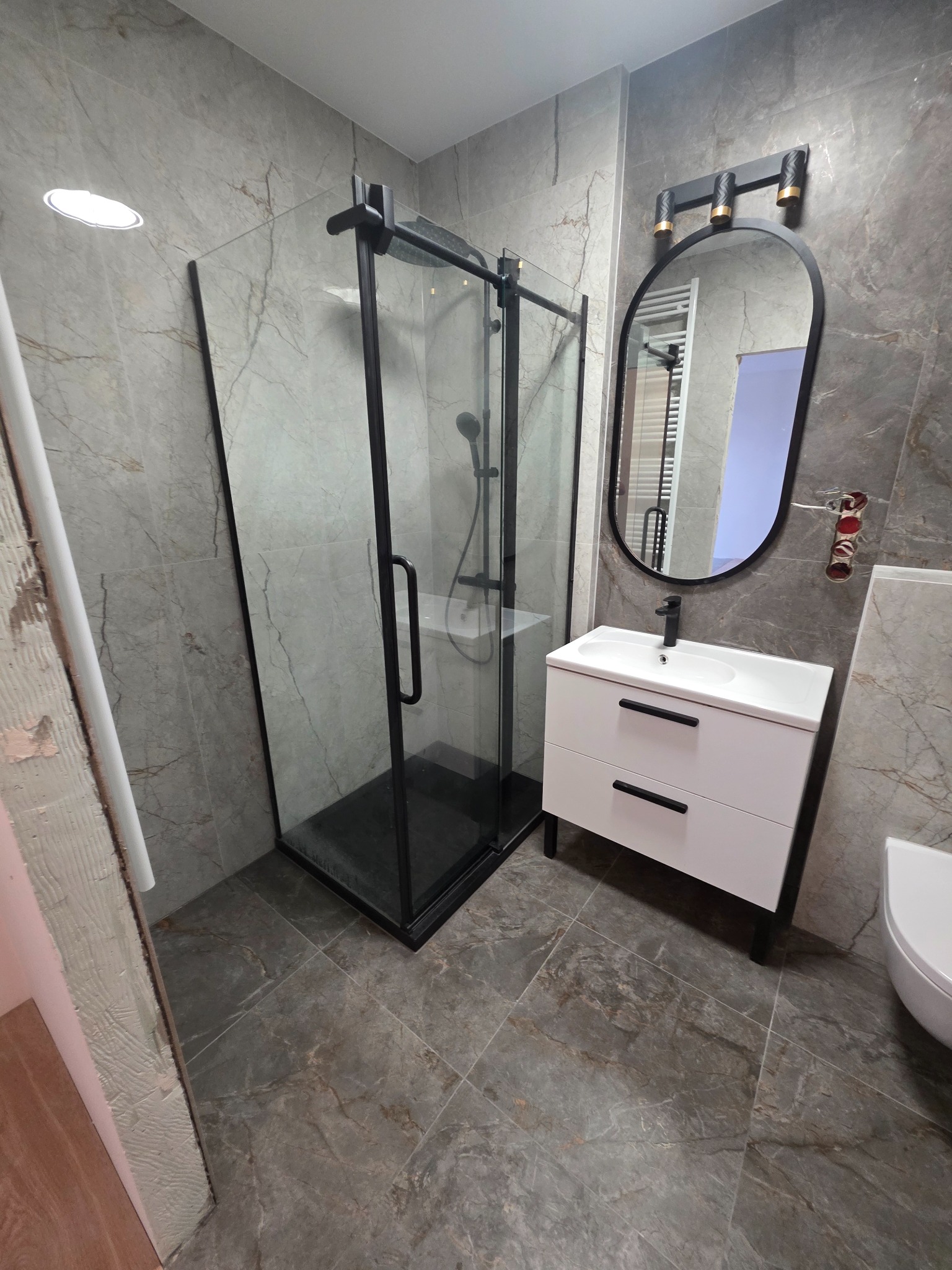 Wykończona łazienka z kabiną prysznicową typu walk-in, umywalką z szafką i lustrem w czarnej ramie. Płytki imitujące marmur na ścianach i podłodze. Widoczne niedokończone wykończenia.