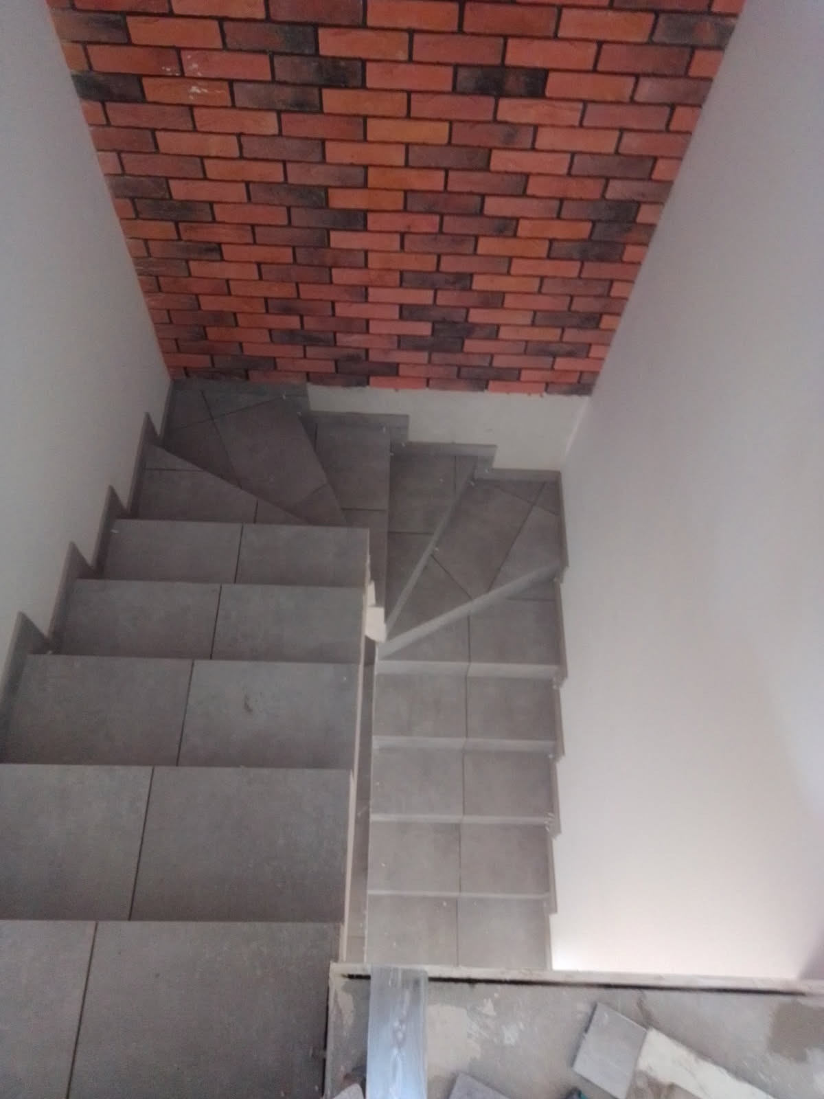 Wykończone schody z szarych płytek, widok z góry. Ściana nad schodami wykończona czerwoną cegłą. Widoczne narzędzia glazurnicze.