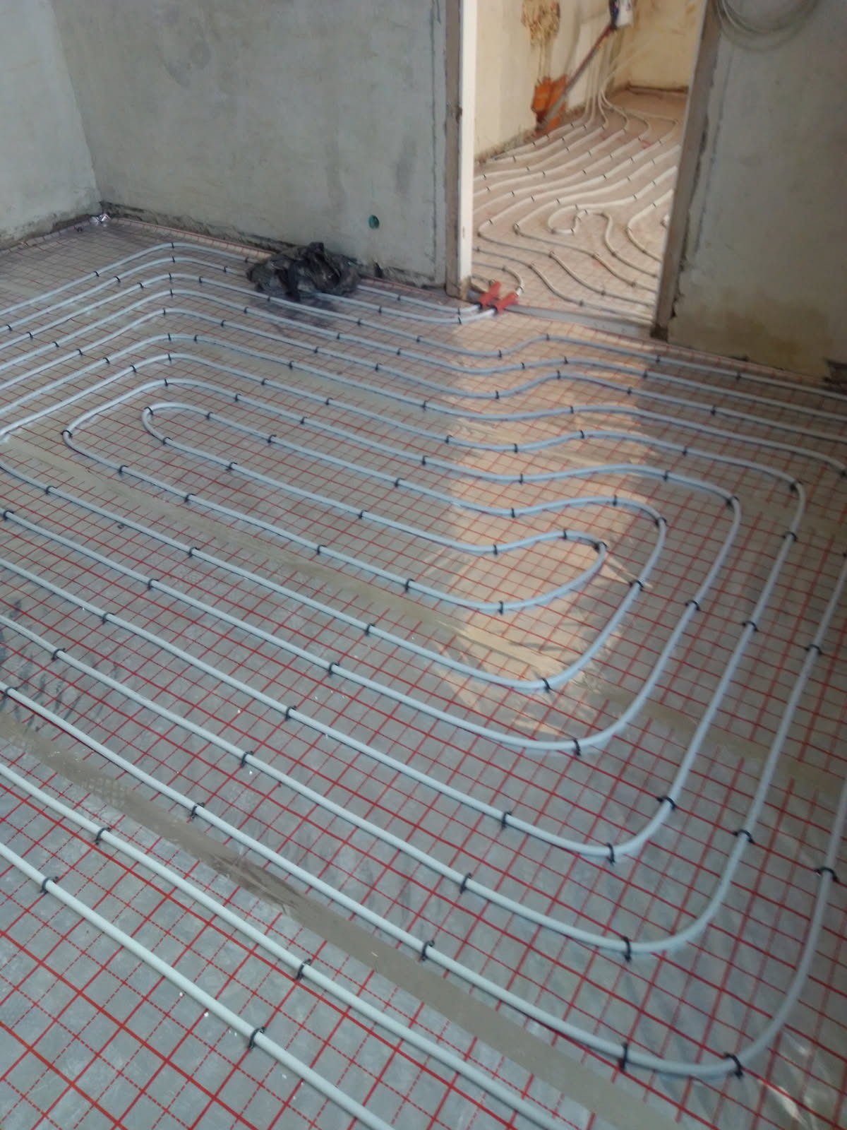 Białe rury ogrzewania podłogowego ułożone na czerwonej siatce w surowym wnętrzu. Widoczne łączenia rur i fragment drugiego pomieszczenia z kontynuacją instalacji.