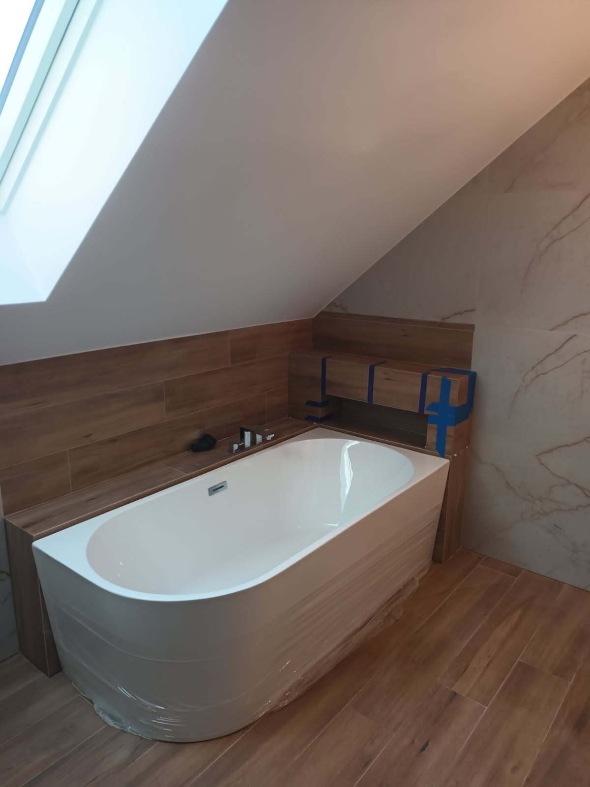 Biała wanna owinięta folią ochronną w łazience na poddaszu z drewnopodobnymi płytkami na ścianach i podłodze. Widoczne okno dachowe.
