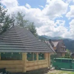 Drewniana altana z dachem pokrytym gontem bitumicznym na tle górskiego krajobrazu i błękitnego nieba z chmurami.