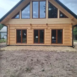 Surowy stan: drewniany dom z bali z ciemnymi oknami i dachem, jeszcze bez zagospodarowania terenu, widok z przodu na tle pochmurnego nieba.