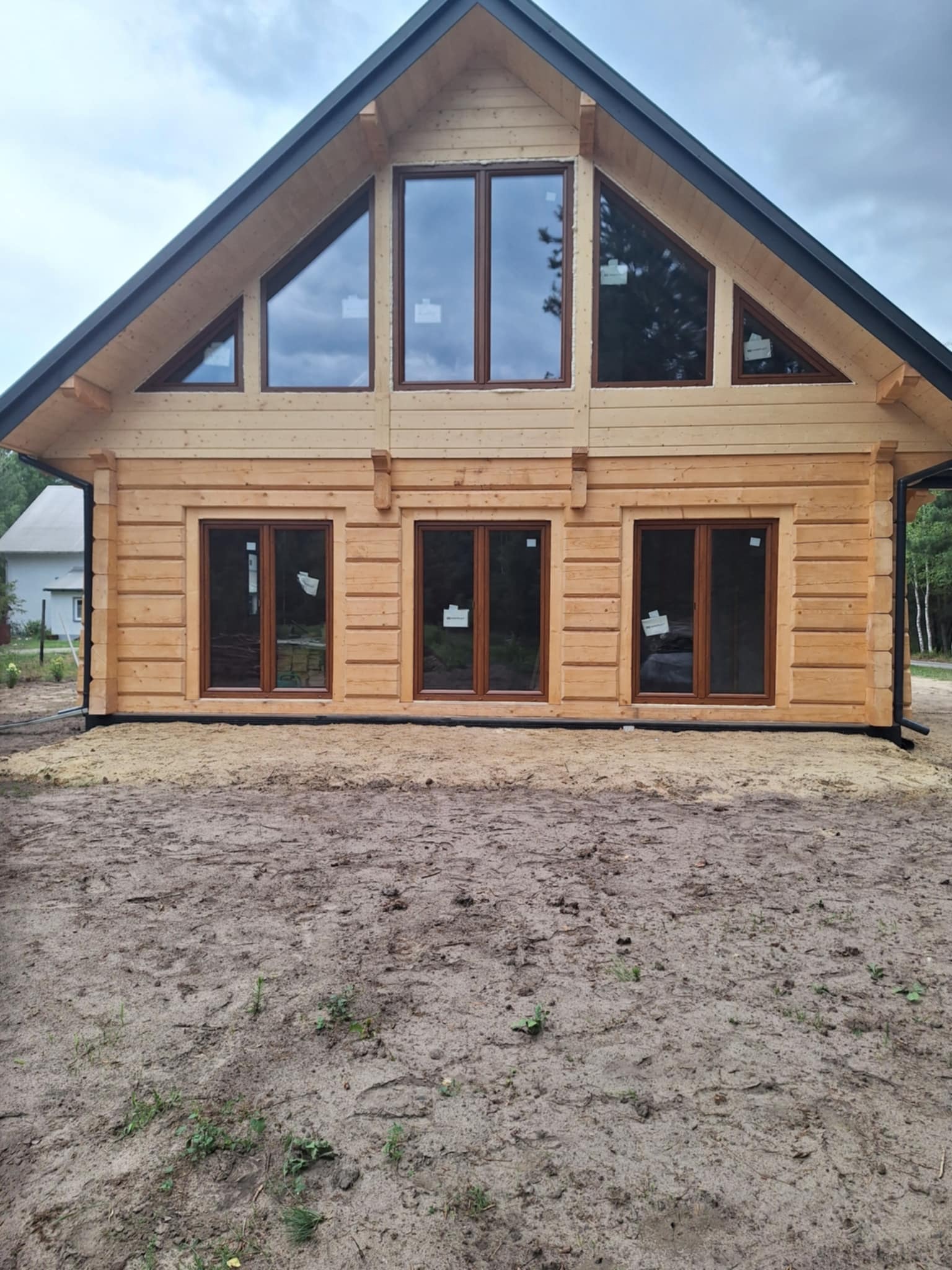 Surowy stan: drewniany dom z bali z ciemnymi oknami i dachem, jeszcze bez zagospodarowania terenu, widok z przodu na tle pochmurnego nieba.