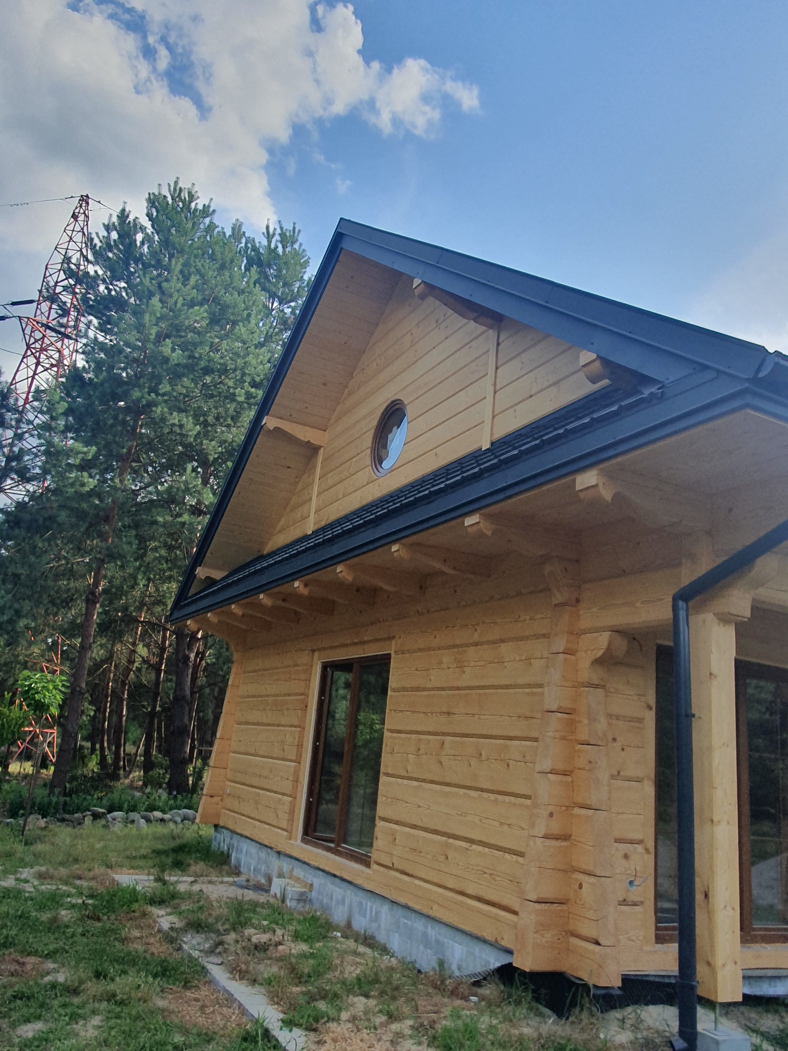 Wykończony dom z bali z okrągłym oknem, ciemnym dachem i widoczną konstrukcją wsporczą w tle, na tle błękitnego nieba z chmurami.